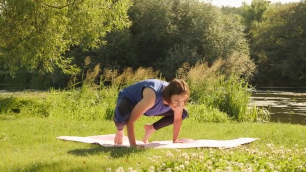 Mujer intenta grúa o cuervo posar y se cae y se ríe. Mujer haciendo yoga en colchoneta de fitness en el parque — Vídeo de stock