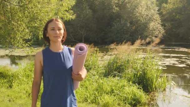 Mujer joven sosteniendo esterilla de yoga para hacer ejercicio al aire libre, caminando en el parque. Vida sana — Vídeo de stock