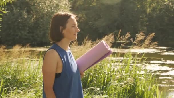 Mujer serena caminando a orillas del río al amanecer. Mujer con esterilla de yoga en las manos que va a practicar yoga — Vídeos de Stock