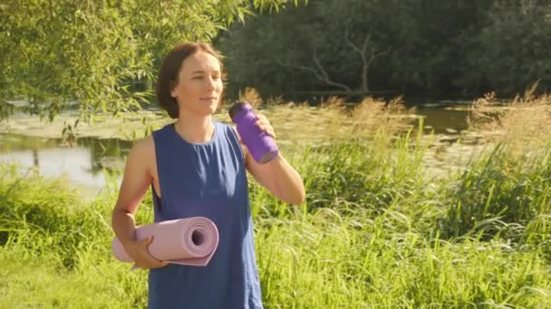 Mujer bebiendo agua, de pie junto al río con esterilla de yoga en las manos por la mañana — Vídeo de stock