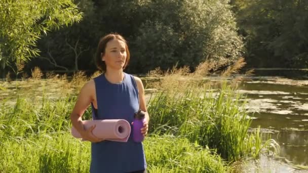 Donna che cammina nel parco dopo la lezione di yoga all'aperto. Femmina con tappetino fitness e bottiglia sportiva passeggiando lungo il lungofiume — Video Stock