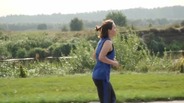 Mujer enérgica corriendo por la carretera a lo largo de la orilla del río. Jogging femenino en el parque — Vídeo de stock