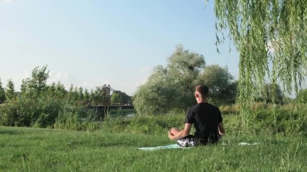 Young man meditating doing yoga exercise by riverside in park in morning — Stock Video