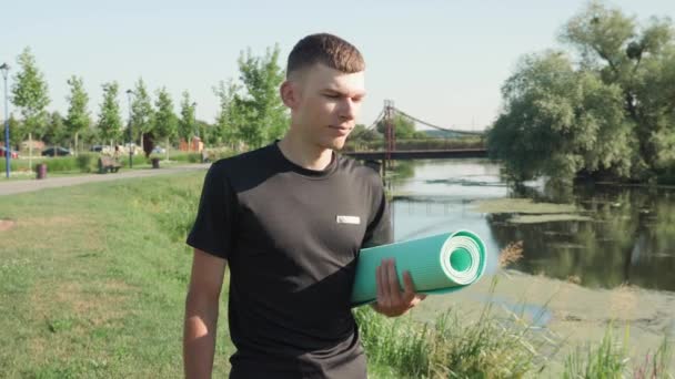 Молодой человек держит коврик для йоги для занятий на открытом воздухе, прогулки в парке. Фитнес-концепция — стоковое видео