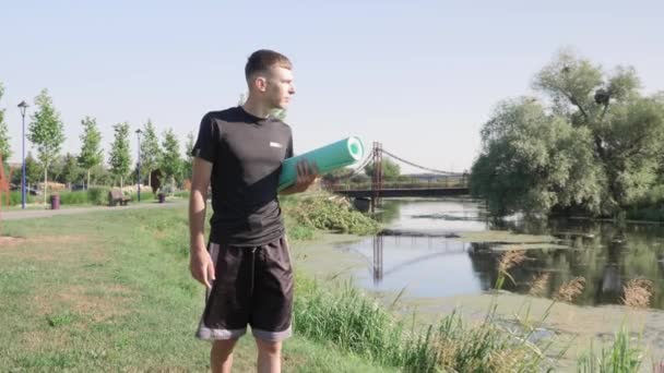Fiatalember sétál a folyó mentén a parkban jóga szőnyeggel a kezében. Férfi jóga edzésre megy. — Stock videók