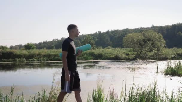 Homme marchant le long de la rivière avec tapis de fitness dans les mains. Homme se promener dans le parc, aller à un cours de yoga — Video