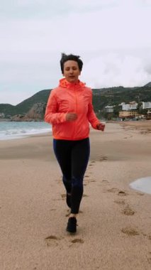 Deniz kıyısındaki kumsalda koşan bir kadının ön loto çekimi. Dişi kardiyo egzersizleri yapıyor, fırtınalı havada kıyı şeridi boyunca koşuyor. Dikey video