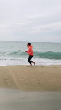 Güçlü genç bir kadın fırtınalı sahilde koşuyor. Kadın koşucu plajda koşuyor. Sağlıklı aktif yaşam tarzı. Spor ve spor konsepti. Dikey Video