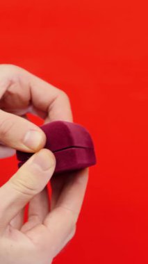 Adam kırmızı arka planda evlenme teklifi için evlilik yüzüğünü kutuda tutuyor. Elleri alyanslı kırmızı kutuyu tutuyor. Sevgililer Günü için hediye. Dikey video