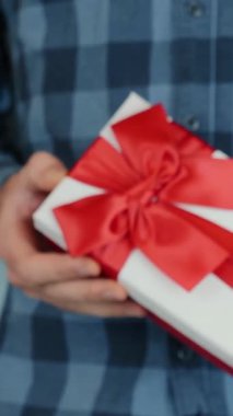 Adam beyaz hediye kutusunu kırmızı yay ile bağlayıp kameraya doğru hareket ediyor. Sevgililer Günü için hediye erkek ellerinde. Dikey video