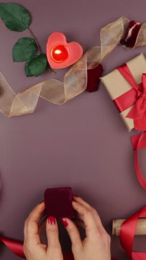 Çiçekler, mumlar, hediyeler ve şömineler eşliğinde Aziz Sevgililer Günü arkaplanındaki nişan yüzüğünün olduğu küçük kırmızı kutuyu eller açıyor. Dikey video