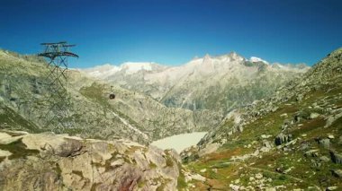Karla kaplı dağ manzarası kayalık dağlar, göl ve hareket eden kırmızı teleferik, hava aracı manzarası. Güneşli yaz gününde İsviçre Alplerinde Grimselsee üzerinde uçan insansız hava aracı