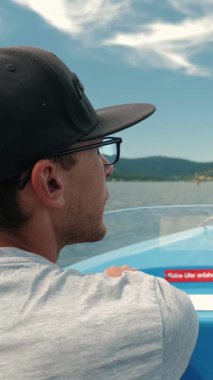 Güneşli bir günde yatta direksiyon sallayan adamın arka görüntüsü. Erkek nehir teknesi, göl boyunca yeşil ağaçlar ve dağlarla çevrili, hafta sonları dinleniyor ve dinleniyor. Dikey video