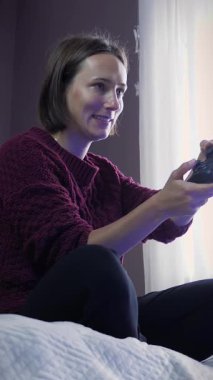Genç bir bayan kanepede oturuyor ve kablosuz joystick kullanarak video oyunları oynuyor. Kadın online oyunlar oynuyor. Dikey video