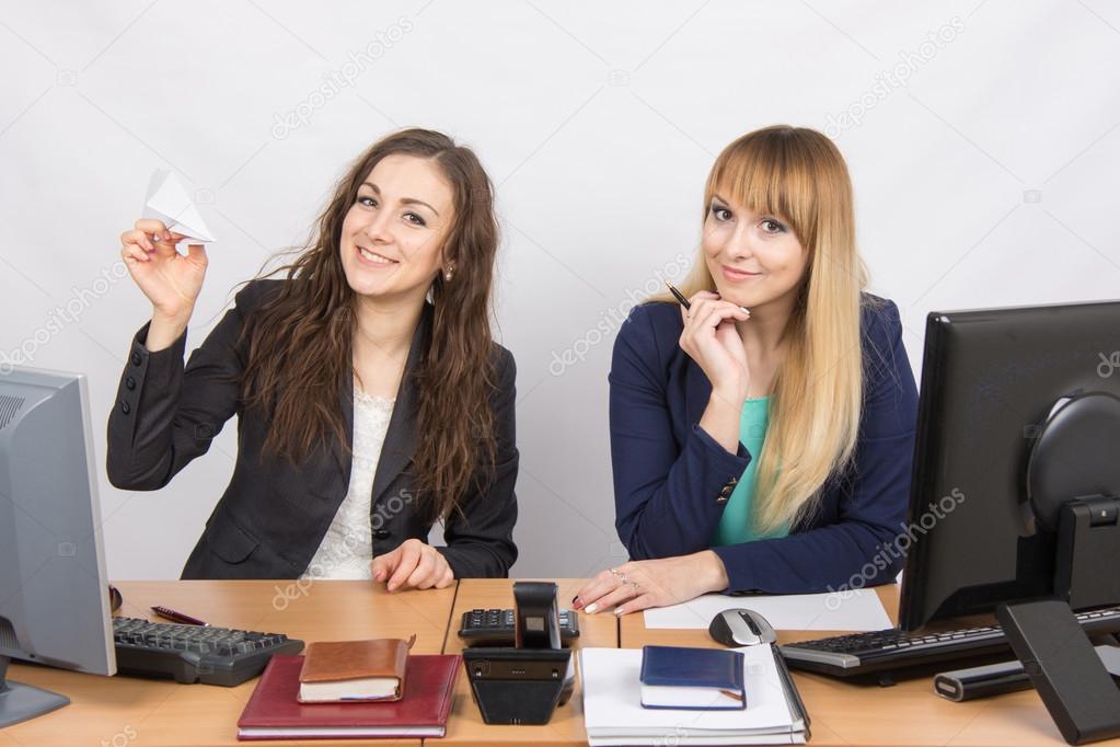Two young women working in the office, one made a paper plane, the second stares into the frame