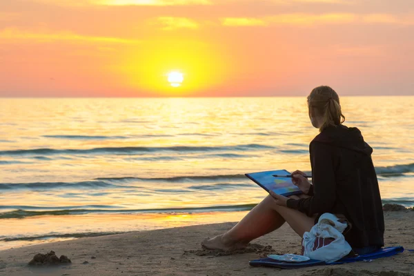 Νεαρό κορίτσι κάθεται στην παραλία, σχέδιο σε ένα φύλλο του ηλιοβασιλέματος ακουαρέλα και κοίταξε τη δύση του ήλιου — Φωτογραφία Αρχείου