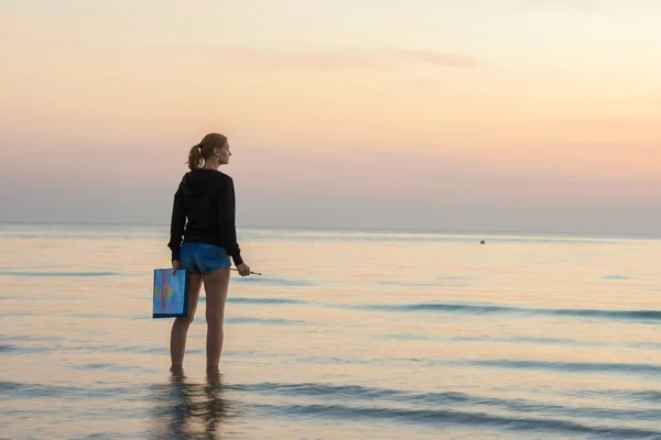 Young girl standing in the water on the beach with a landscape painted on a sheet of paper and brush — Stock Photo, Image