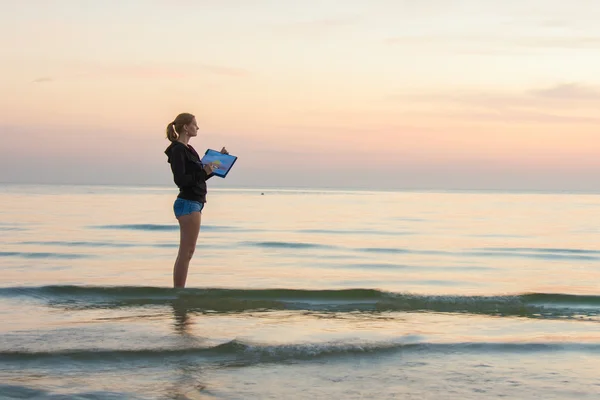 Genç kız sahilde suyun içinde duruyor ve günbatımının renklerini bir kağıt parçasına çiziyor. — Stok fotoğraf