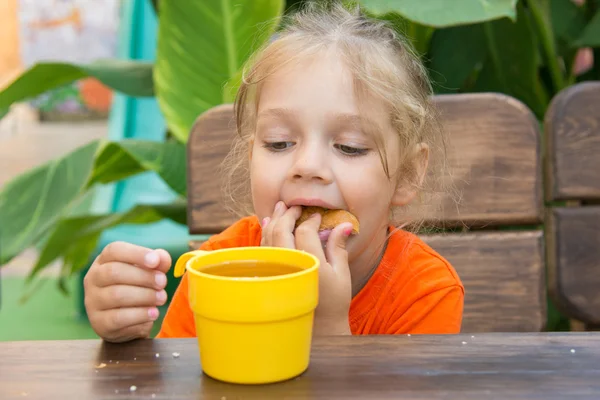 Menina de quatro anos engraçado recheado bun em sua boca e olha para um copo de chá — Fotografia de Stock