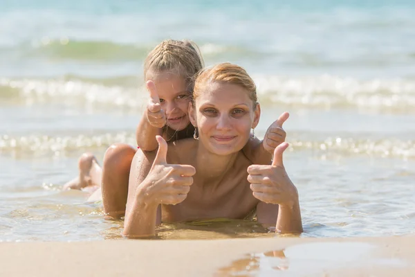 Mum with a daughter on his back lying on the beach and show. Girl and woman show thumbs up — Stock Photo, Image