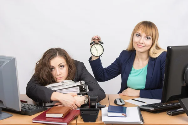 Dívka v kanceláři s úsměvem ukazuje čas na hodinách vedle vyčerpaného kolegy — Stock fotografie