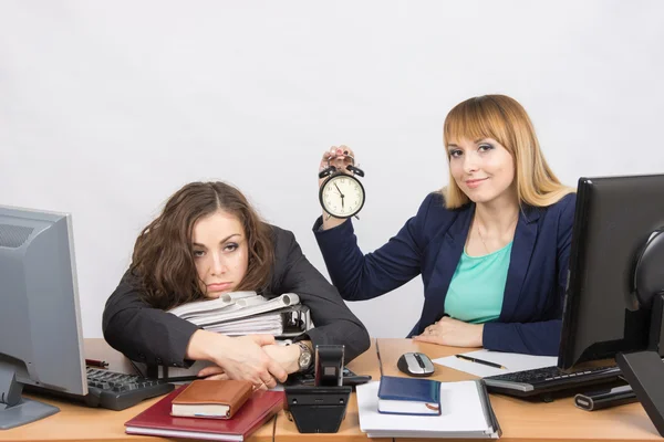 Dos chicas en la oficina al final del día, una con una sonrisa, sosteniendo un reloj, otra cansada yace en carpetas —  Fotos de Stock