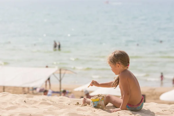 Пятилетняя девочка, сидящая на пляже и играющая с песком и игрушками на фоне моря — стоковое фото