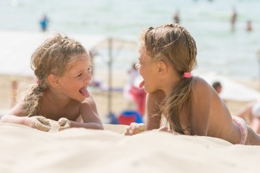 Two happy little girls show each other tongues on a sandy beach clipart