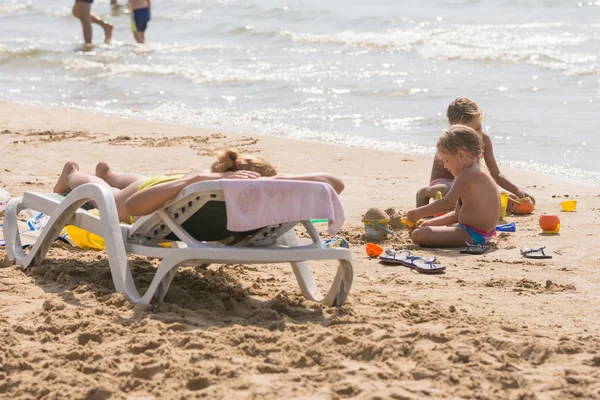 На морском пляже рядом с водой с шезлонгом для загара Женщина и дети играют на песке — стоковое фото