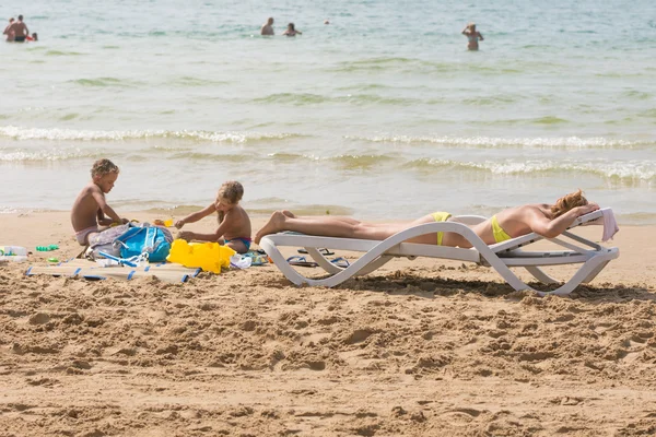 En la playa cerca del agua con una silla de playa tomando el sol mujer cerca de los niños jugando en la arena — Foto de Stock