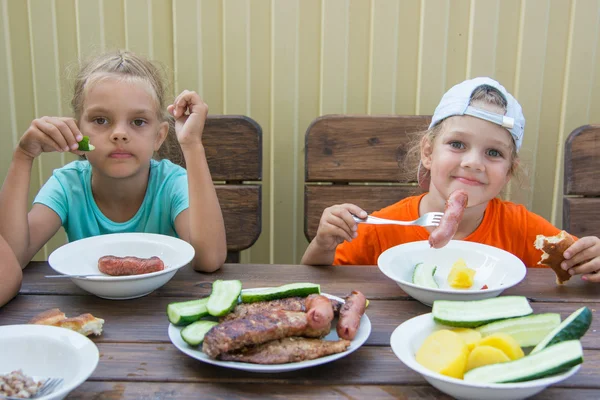 Two little girls at a wooden table in nature eating grilled sausages — Stock Photo, Image