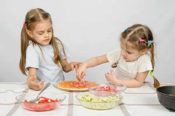 Šestiletá dívka sleduje a ovládá svou mladší sestru umísťuje ingredience na pizzu — Stock fotografie
