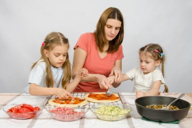Anne küçük kızı bir pizza ketçap yaymak yardımcı olur, büyük kızı, o ikinci bir pizza hazırlanıyor