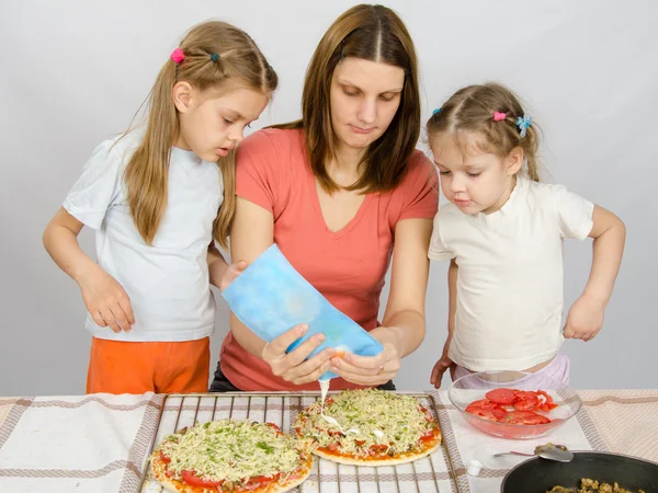 Две маленькие девочки смотрят с интересом, как мать поливает майонезную пиццу — стоковое фото