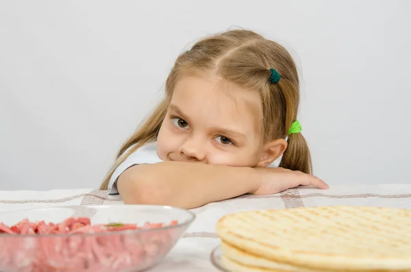 Liten flicka sitter vid ett bord med huvudet på sin hand med ett leende och titta på maten framför henne — Stockfoto