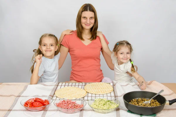 Mother with two daughters at the kitchen table are going to cook a pizza and a fun look into the frame — Stock Photo, Image