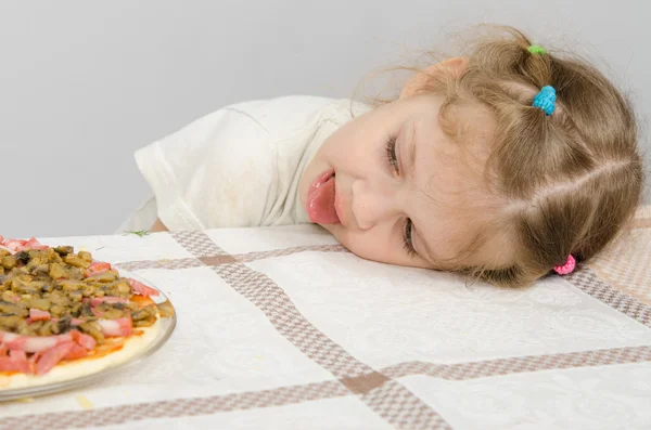 Kleines Mädchen mit herausgestreckter Zunge legt den Kopf auf den Tisch und blickt auf die Pizza — Stockfoto