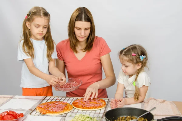 Den äldsta dottern hjälper mamma laga en pizza, och den yngsta är att titta på dem — Stockfoto