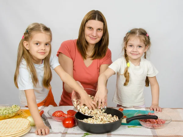 Two little daughter at the kitchen table helping her mother pour chopped mushrooms from the plate to the pan — Stock Photo, Image