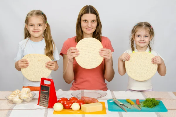 Mamá con dos niñas sentadas en fila en la mesa de la cocina y unas pizzas de mano —  Fotos de Stock