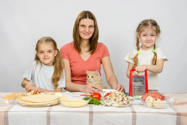 Mor och två små flickor vid ett bord förberedda ingredienser för pizza. De såg en katt — Stockfoto