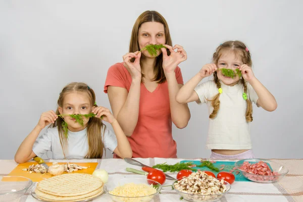 Jovem dona de casa com duas filhas se divertindo segurando raminho de salsa como um bigode na mesa da cozinha ao compartilhar cozinhar — Fotografia de Stock