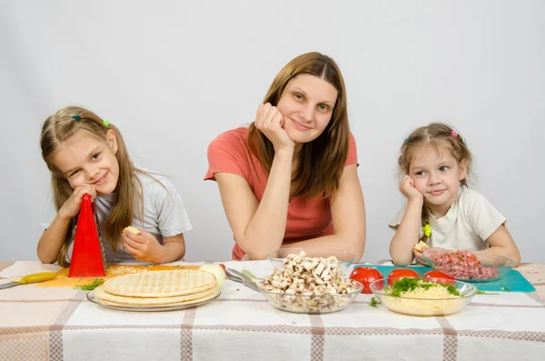 Satisfeito mãe com duas filhas sentadas descansando a cabeça em suas mãos na mesa com os produtos para pizza — Fotografia de Stock