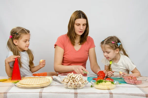 Mamma visar två unga döttrar vid köksbordet som skär tomat pizza — Stockfoto