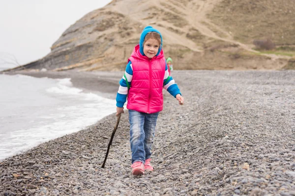 Пятилетняя девочка ходит по галечному пляжу в теплой яркой одежде с палкой в руке — стоковое фото