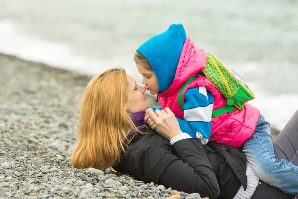 5-jähriges Mädchen sitzt auf der Seite einer Mutter, die am Kiesstrand liegt und küsst sie auf die Nase — Stockfoto