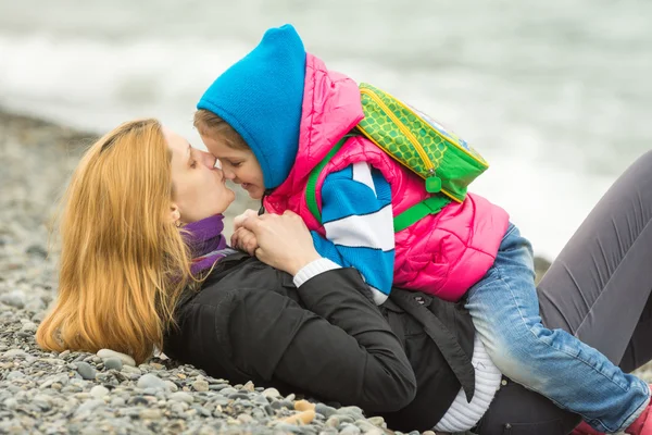 Junge Mutter liegt auf dem Kieselstrand und küsst die Nase ihrer Tochter, die auf ihrem Reiten sitzt — Stockfoto