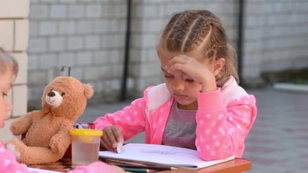 Семилетняя девочка забавно стирает ластик, рисуя с сестрой на улице — стоковое видео