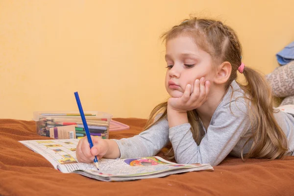 Bambina che studia una rivista con una matita in mano sdraiata sul suo stomaco e la testa nella sua seconda mano — Foto Stock