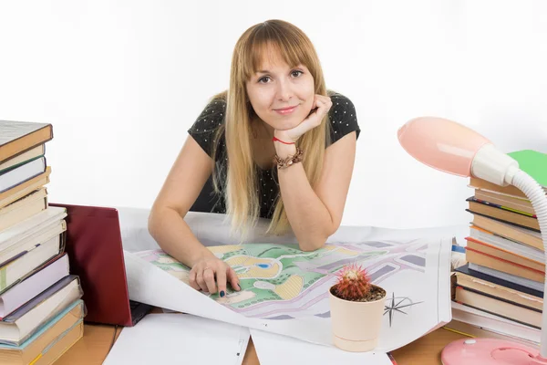 De student van een meisje aan een bureau vol met boeken en tekeningen met een glimlach ziet er in het frame — Stockfoto